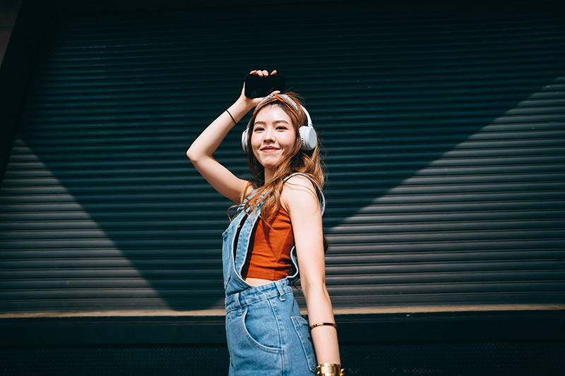 Portrait of carefree, smiling young Asian woman holding smartphone, dancing while listening to music on headphones outdoors against coloured wall and sunlight. Music and lifestyle