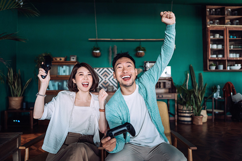 Happy young Asian couple sitting on the sofa in the living room, shouting in excitement while having fun playing video games together at home
